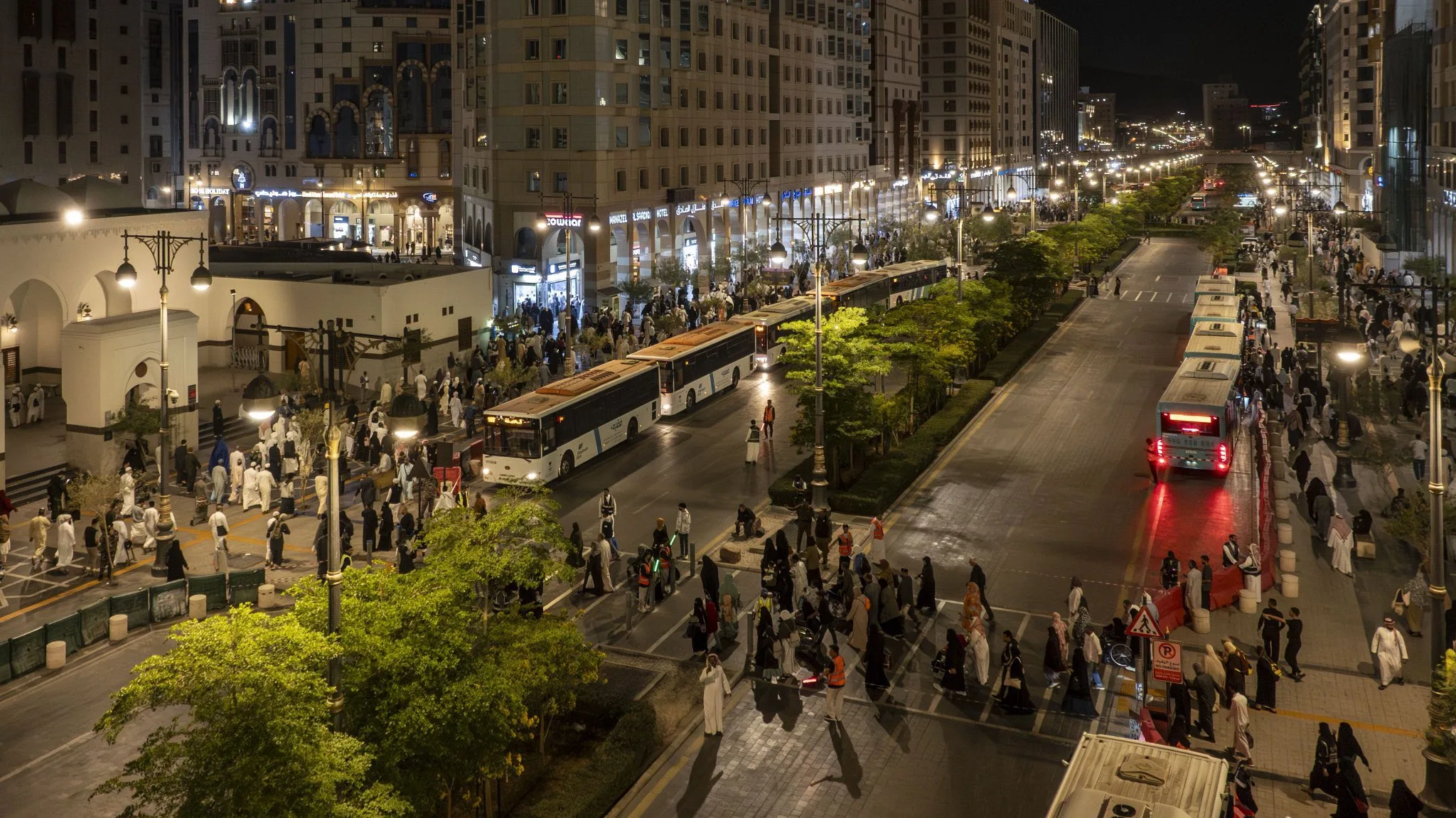 Nighttime city street crowded with pedestrians, buses, and bright storefronts along a tree-lined avenue.