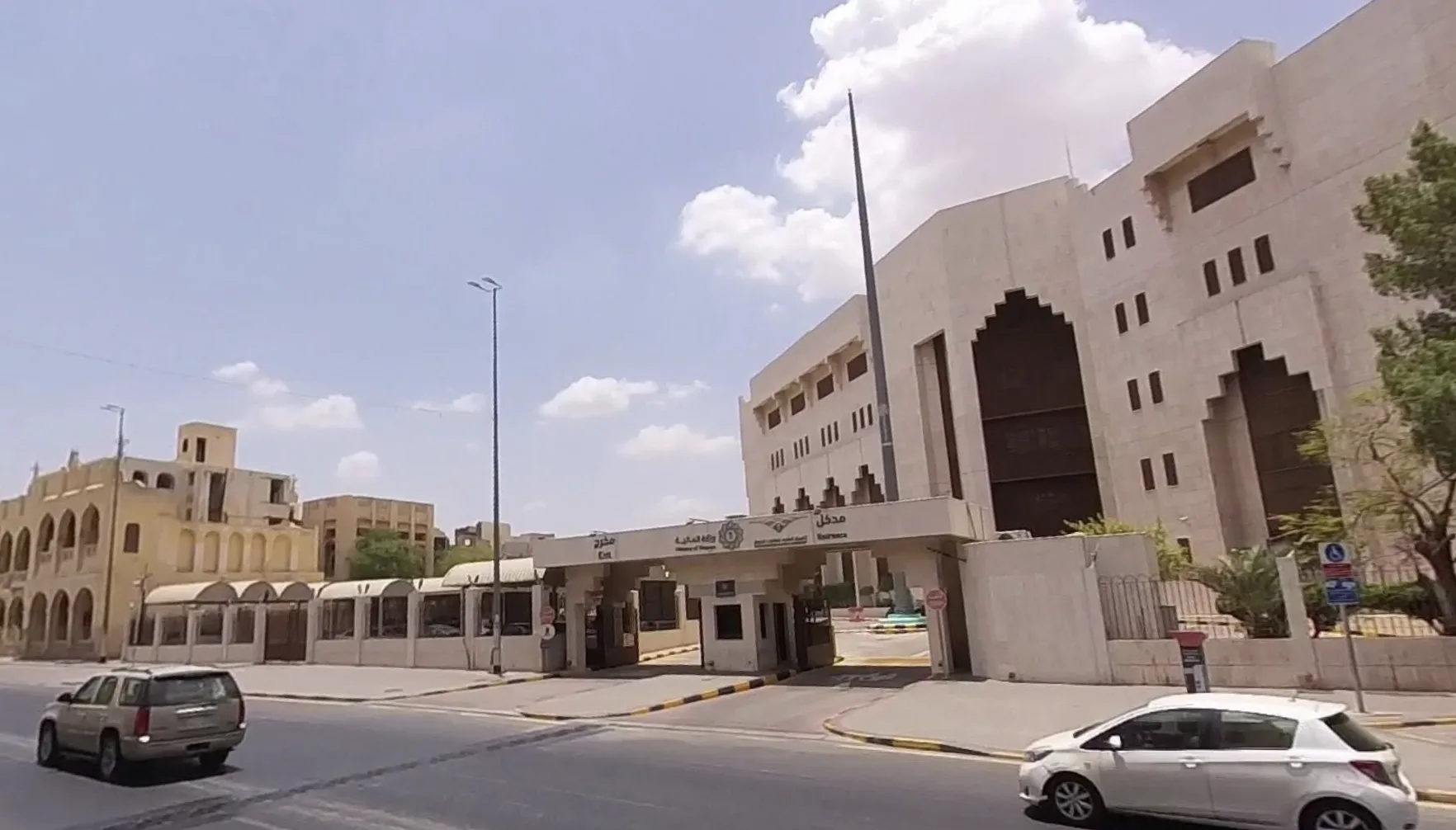 Wide street in front of a beige institutional building with a large arched entrance and gated driveway, under a blue sky with clouds, and a few cars.