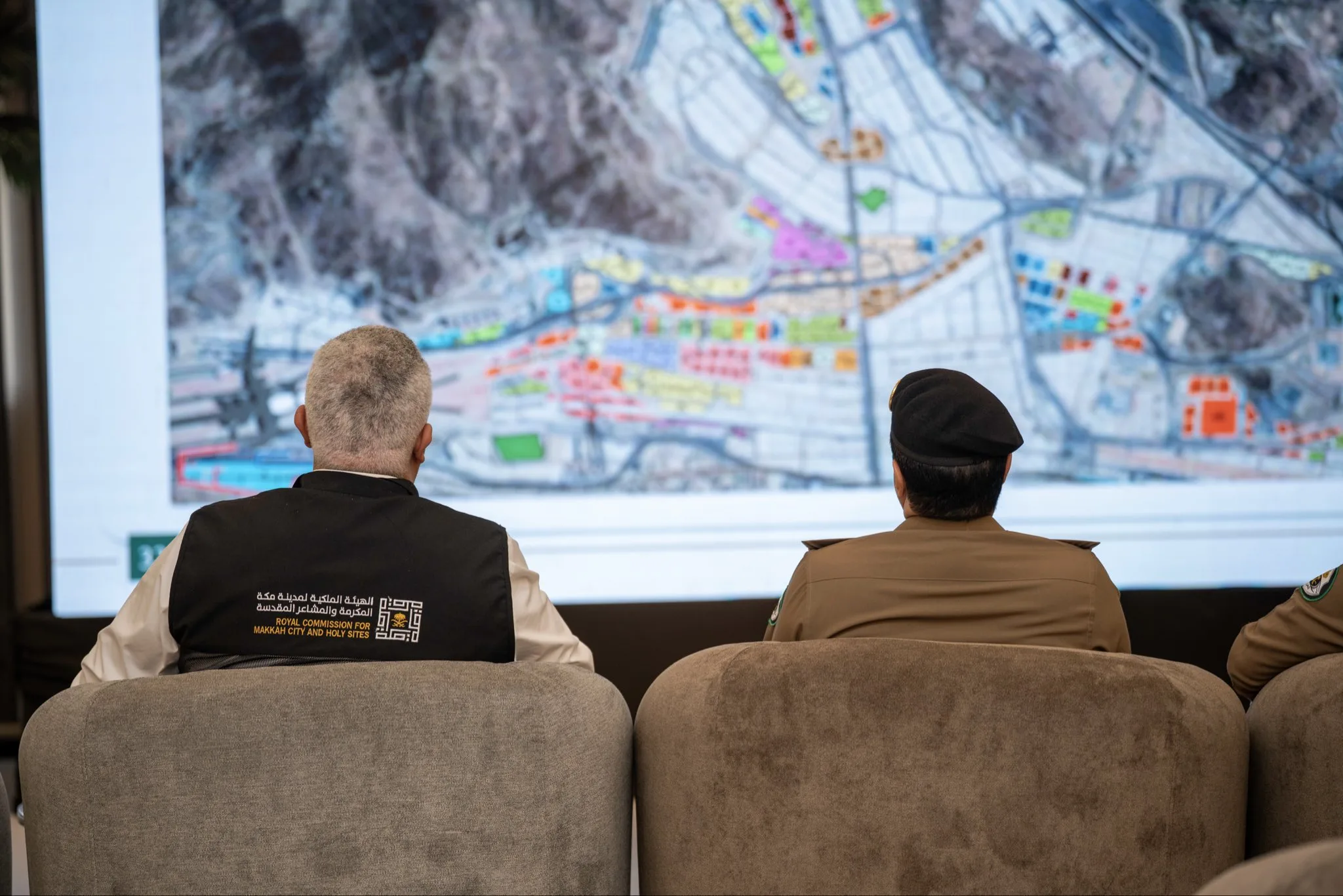 Two men in uniforms sit facing a large, colorful map on a wall, viewed from behind.
