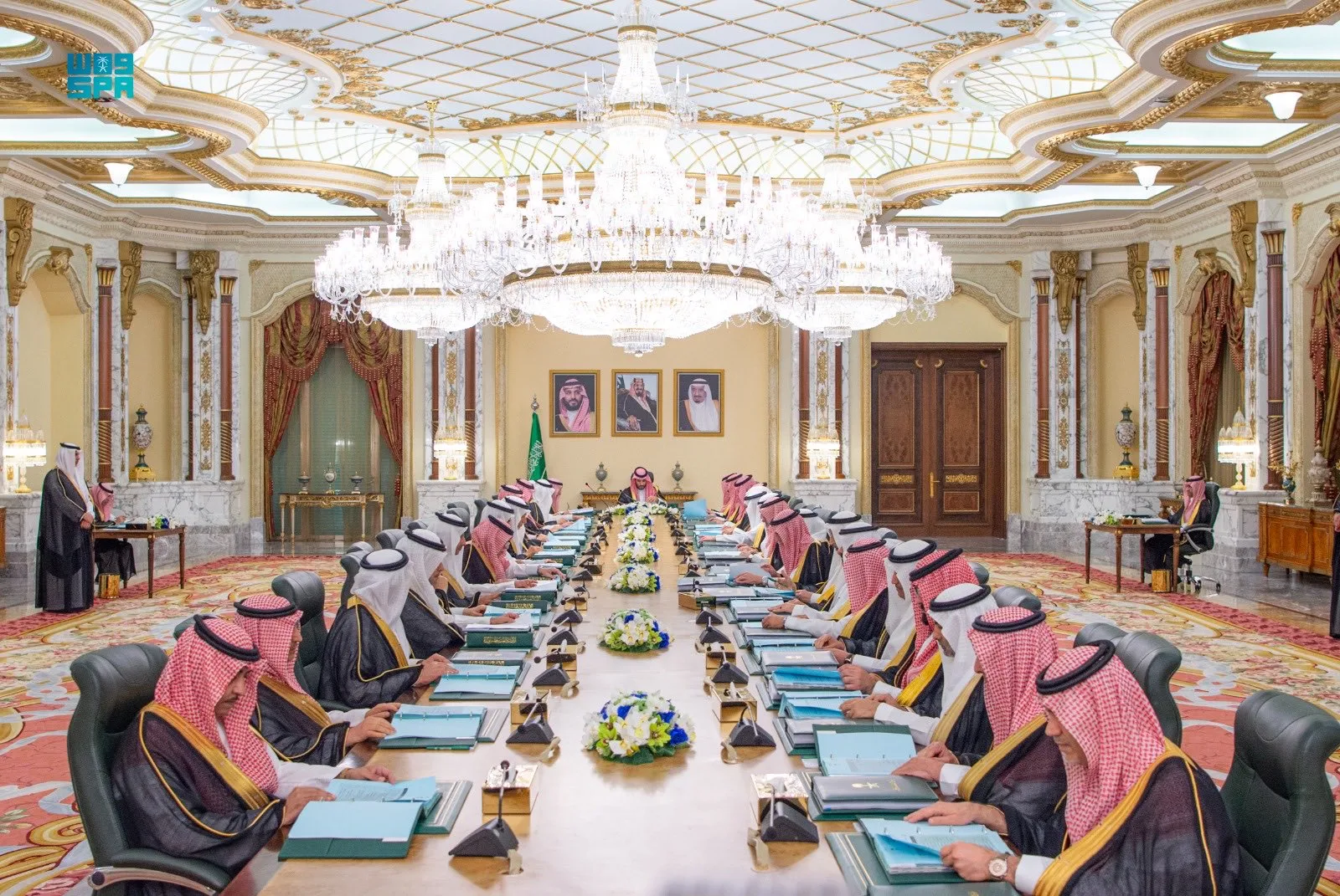 Formal Saudi meeting in an opulent room with a long table, officials in traditional attire, and a crystal chandelier overhead.