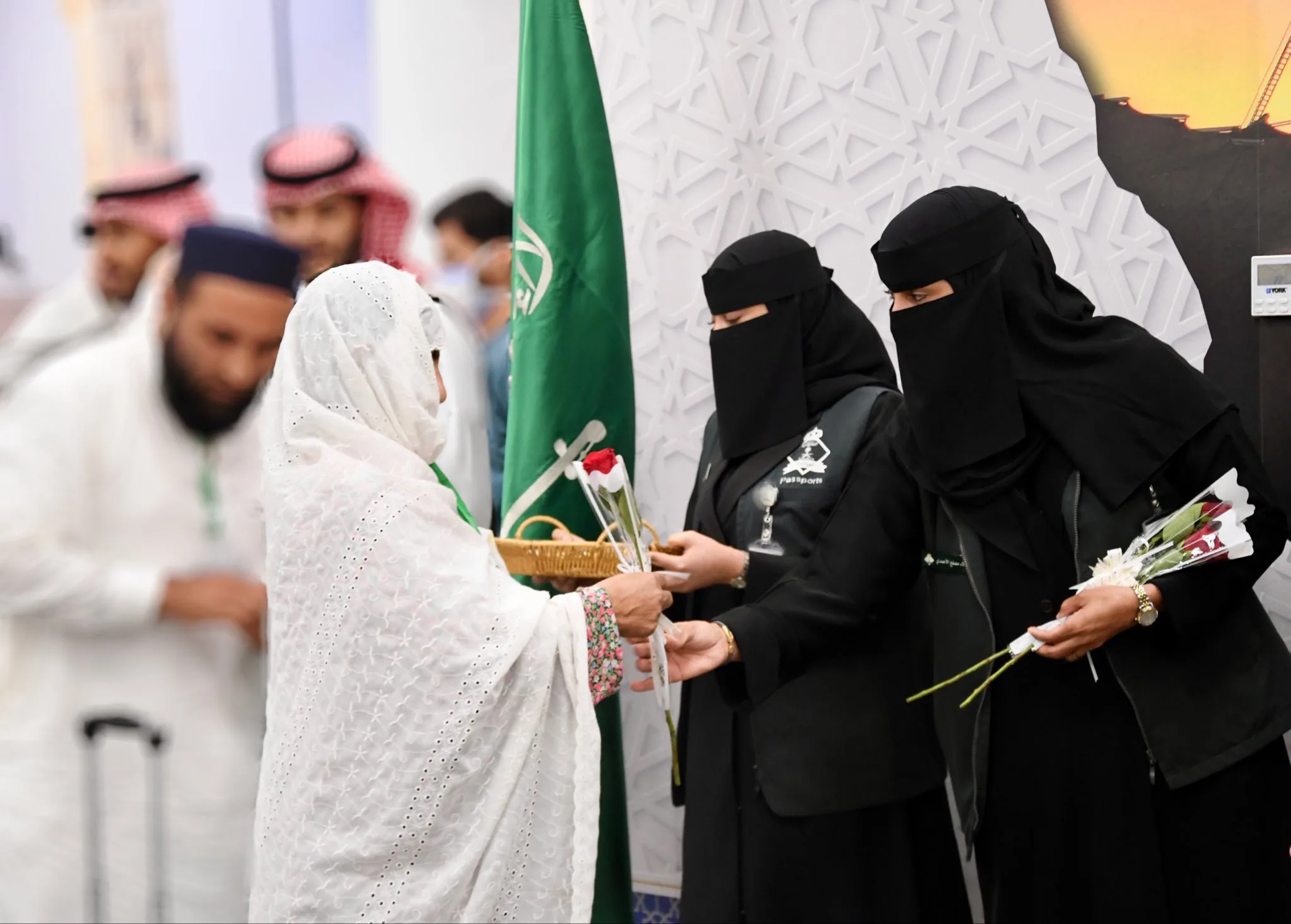 Woman in a white traditional dress hands a red rose to two women in black niqab at a ceremony, with a green flag and patterned wall in the background.