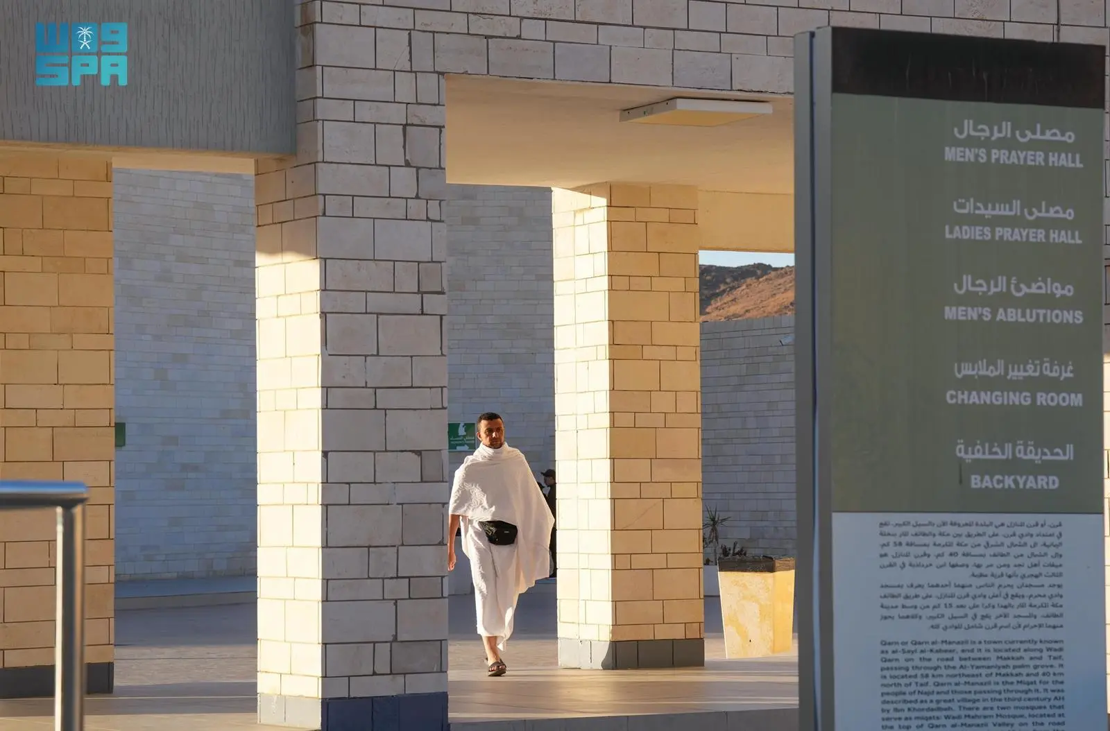 Man in white robes walking through a stone colonnade at a religious site, with a tall sign listing prayer halls in the foreground.