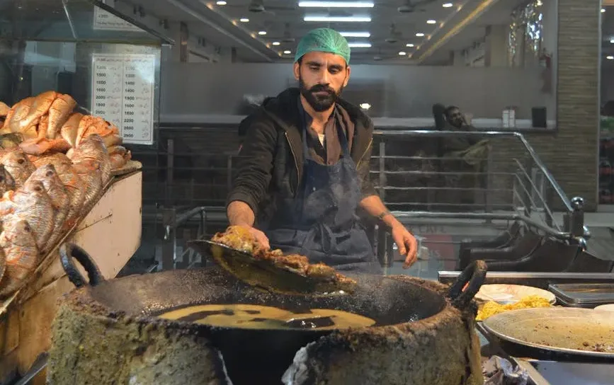 Male cook in a teal hairnet and apron flips meat on a large skillet at a market stall, with loaves of bread stacked to the left.