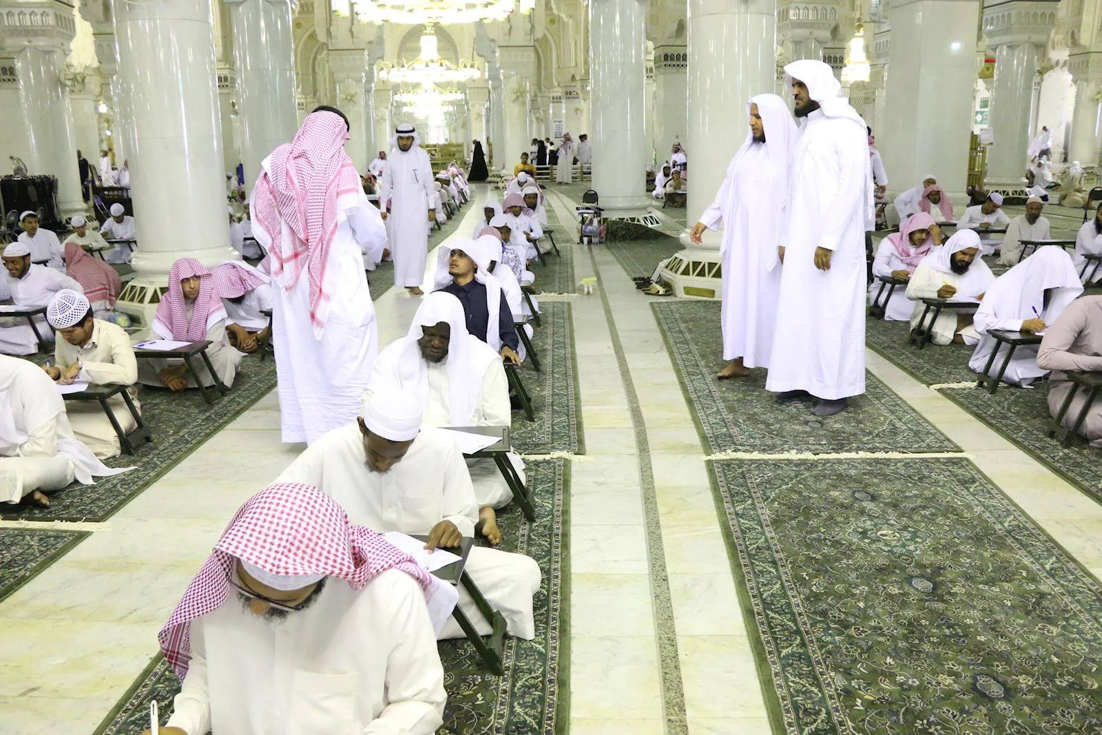 Men in white robes sit on the floor, writing at portable desks inside a grand, columned mosque hall with green carpets.