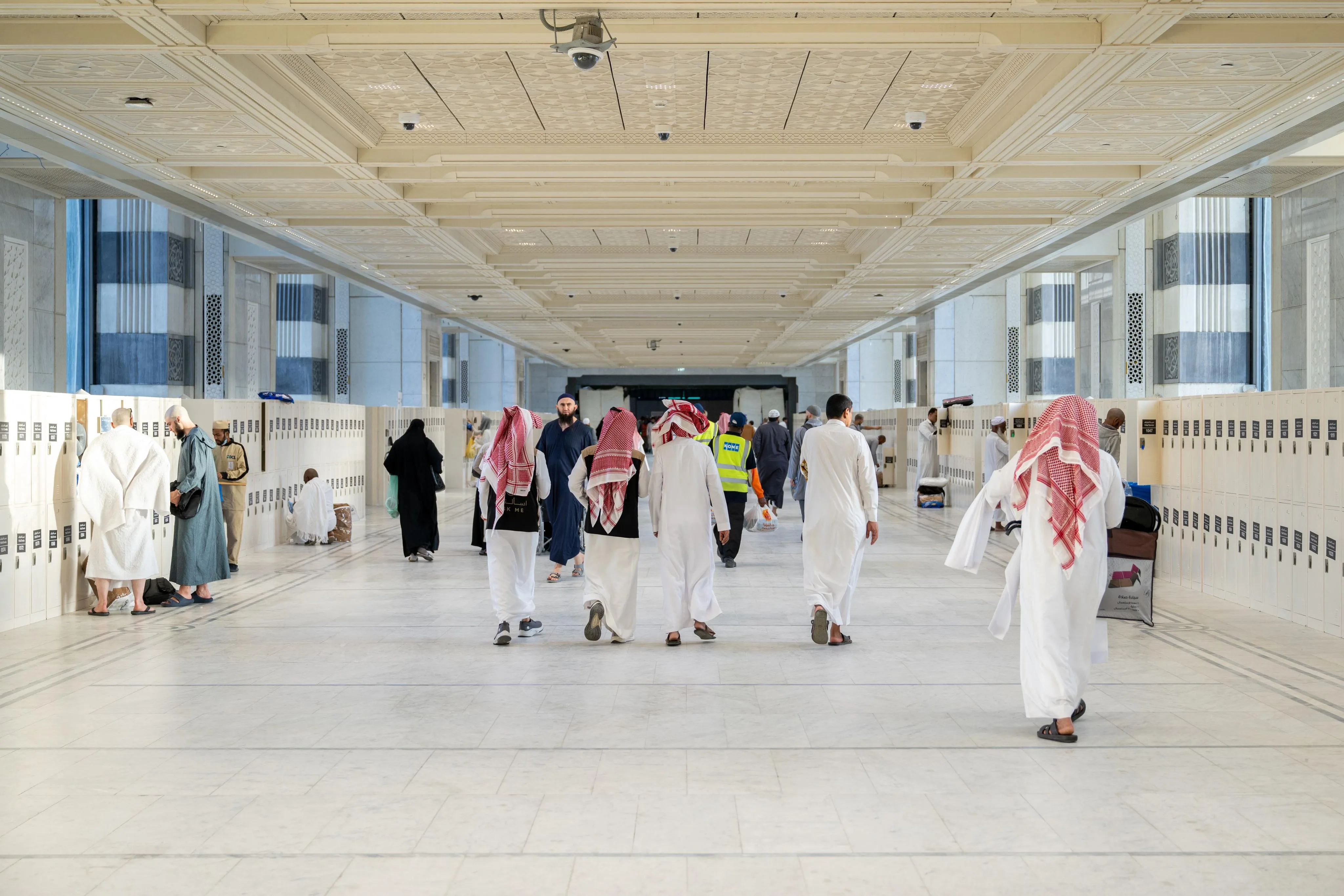 Busy marble corridor filled with pilgrims in white ihram and traditional clothing, walking between rows of lockers on both sides under a detailed ceiling with security cameras overhead