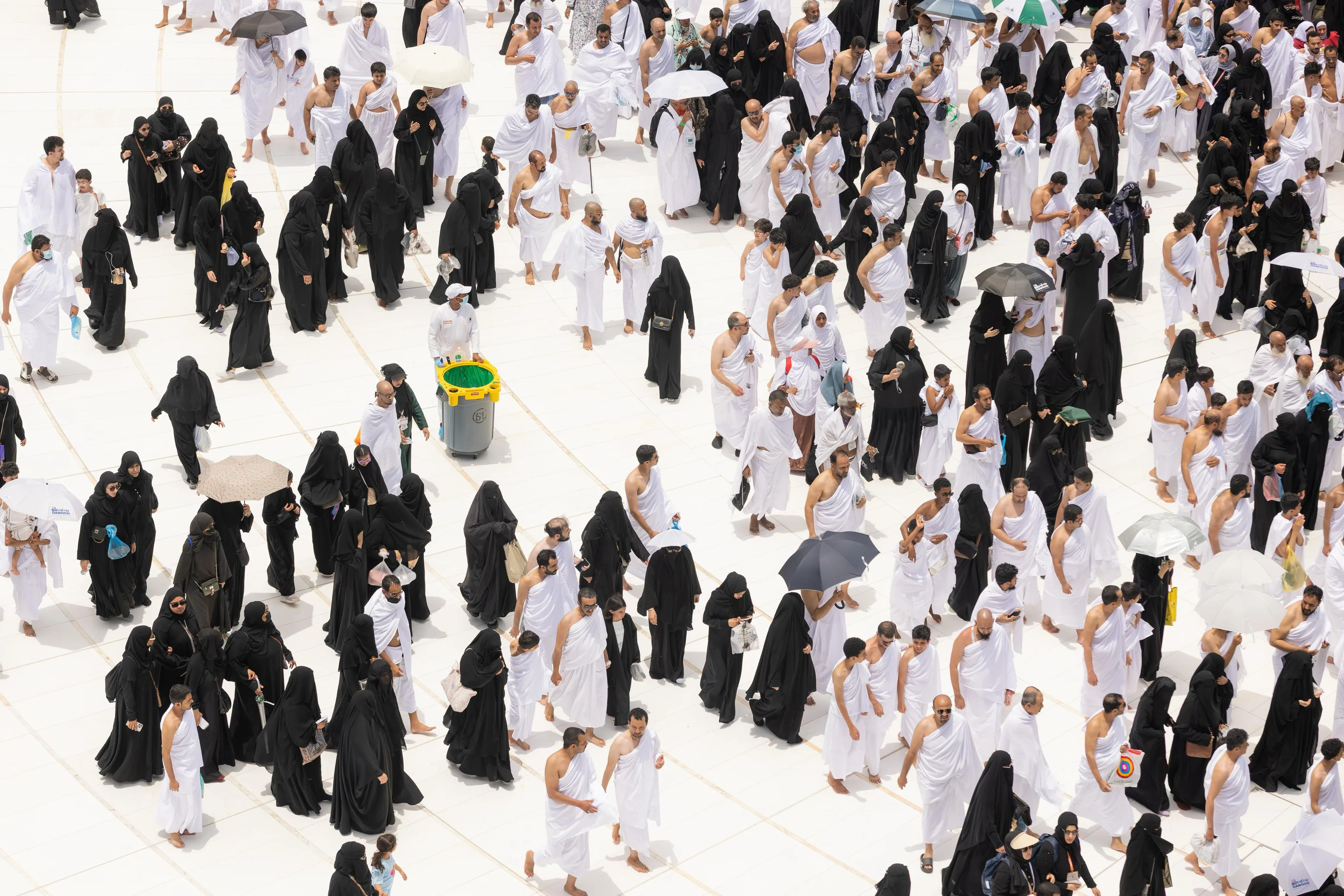 Aerial view of a large crowd of Muslim pilgrims in white ihram and black abayas walking on a white tiled plaza.