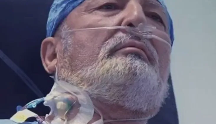 A close-up of a male patient in surgery with an oxygen cannula and blue surgical cap, looking upward.