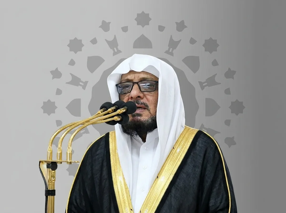 Religious leader in white head covering and black and gold robe speaks into multiple microphones.Provided in a ceremonial setting with a decorative backdrop.