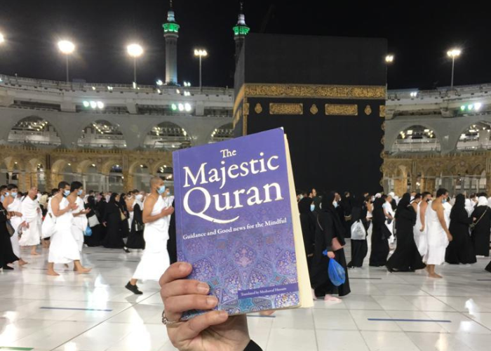 The Sacred Journey How Hajj and the Quran Are Inseparably Linked - Nayi Soch News-e-Hub Person holding a purple copy of 'The Majestic Quran' in front of the Kaaba during Hajj, with pilgrims in white and black robes around. Substantial sacred backdrop.