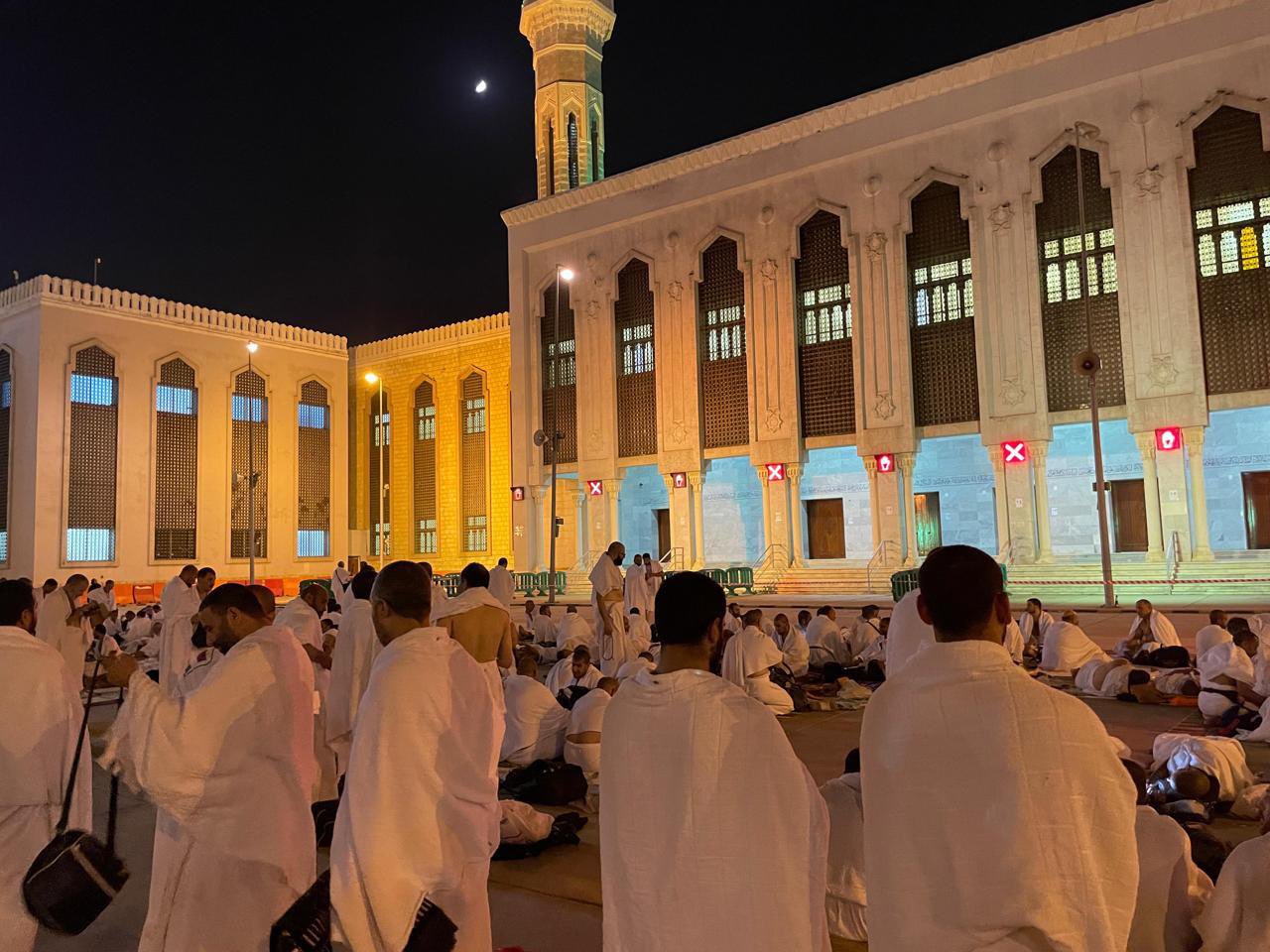 Group of Muslims in white ihram robes sitting on a mosque courtyard at night with a tall minaret in the background.