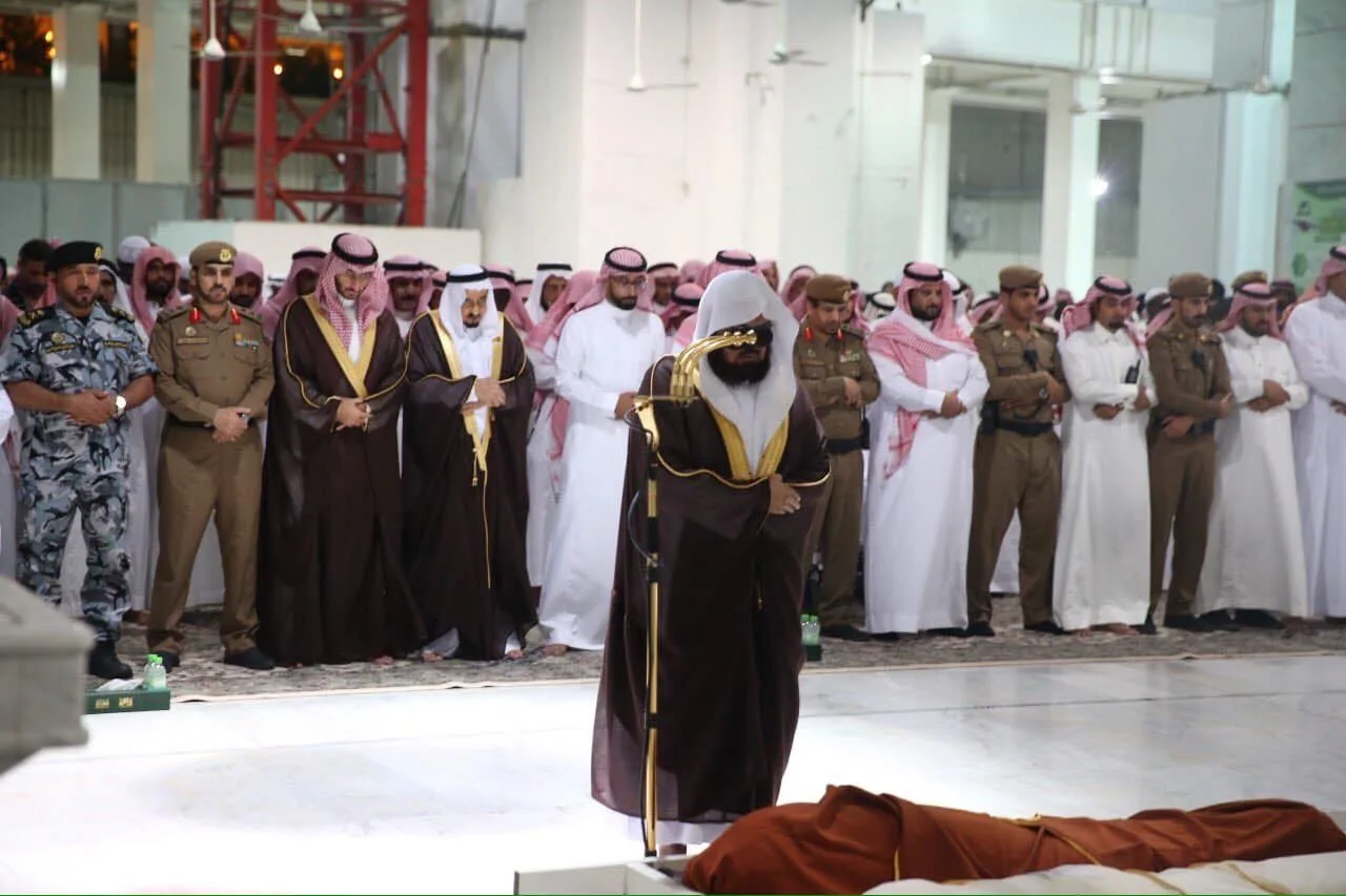 Imam in brown robes leads a funeral prayer as a crowd of men in traditional Saudi attire and uniforms stand behind him, with a wrapped body in front.