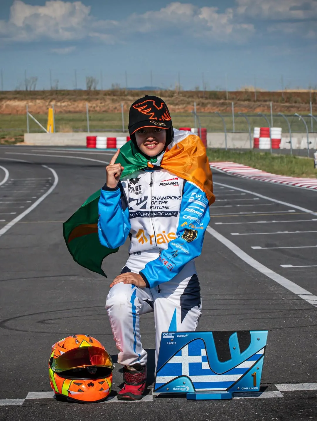 Young go-kart racer on a track, wearing a blue racing suit and an orange-green cape, kneeling with an orange helmet beside them and a blue race plaque nearby.