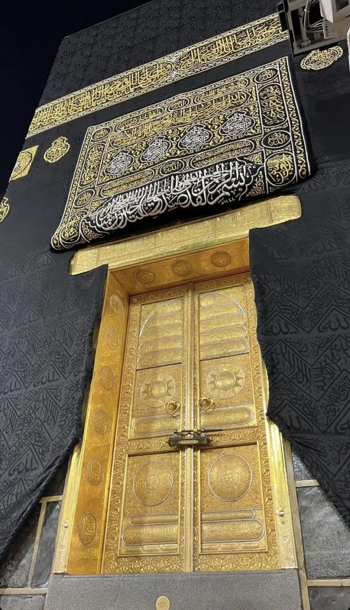 Gold-plated Kaaba door with ornate panels, set in a black Kiswah adorned with gold Arabic inscriptions at night