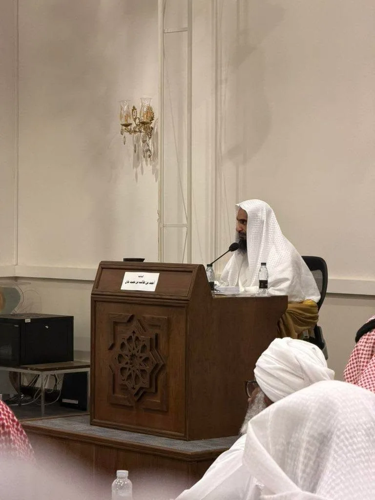 Man in white traditional attire speaks at a wooden podium with a microphone in a conference room.