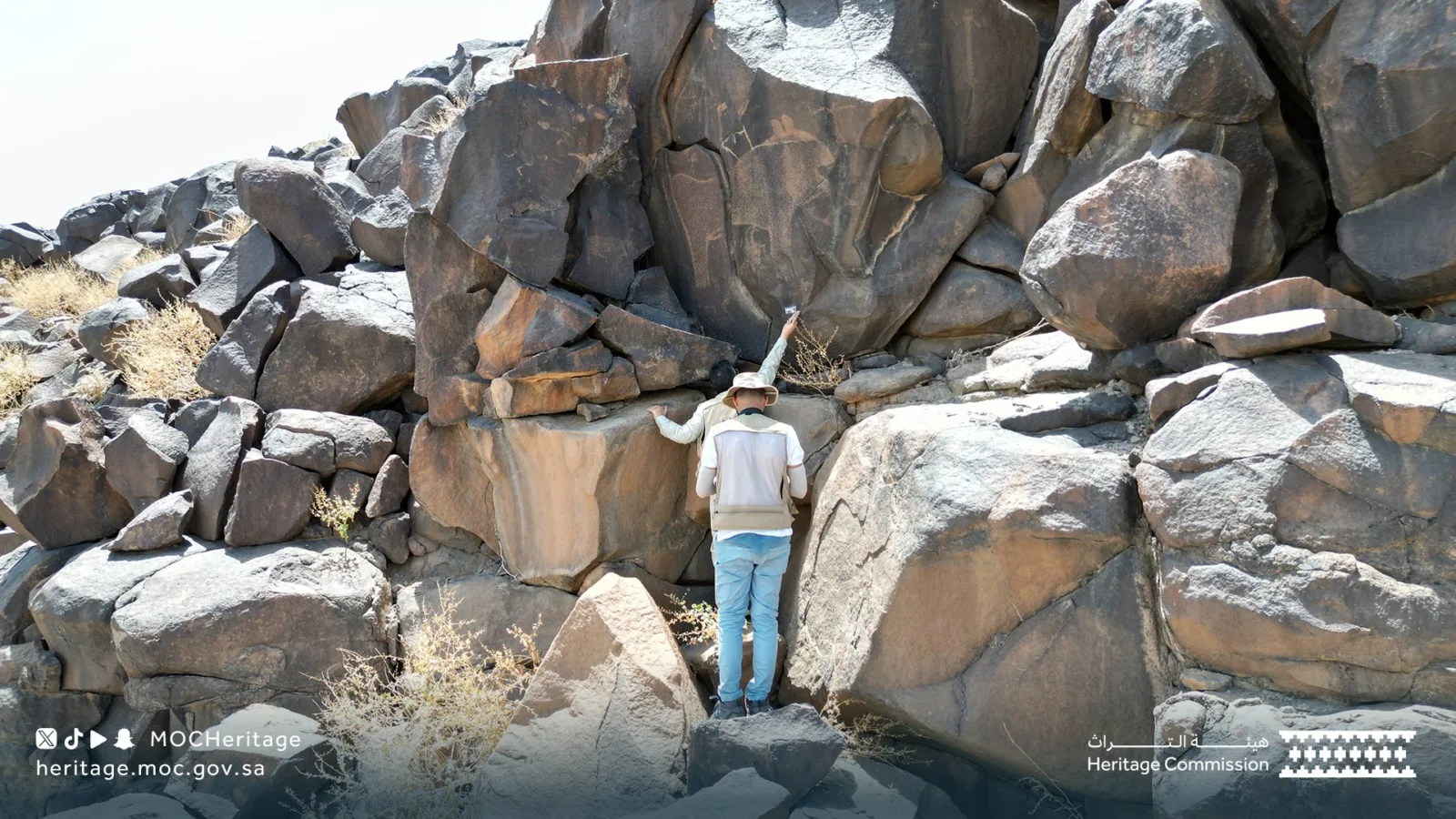 Rare Ancient Rock Text Mentioning Umar RA Found in Madinah - Nayi Soch News-e-Hub Person wearing a light vest and hat stands among large desert rocks, reaching up to touch a boulder while exploring a rocky hillside. Dry shrubs dot the landscape; heritage logos appear in the lower corners.