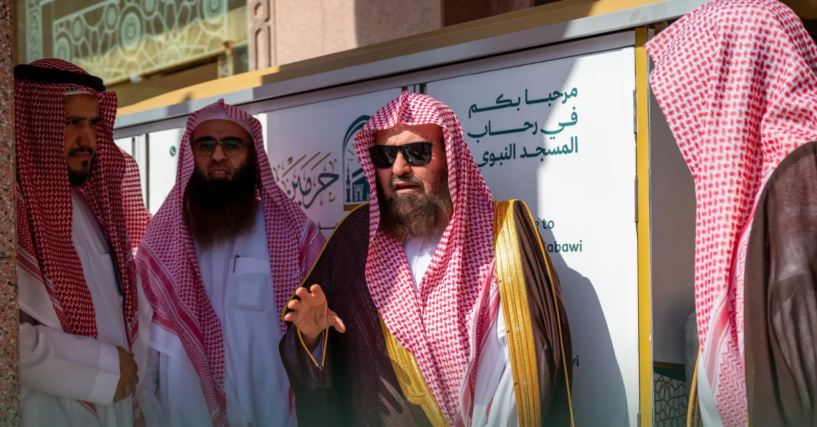 A bearded man wearing a red-and-white keffiyeh and dark robes speaks while gesturing, flanked by two other men in traditional attire against a white sign with Arabic text in the background.