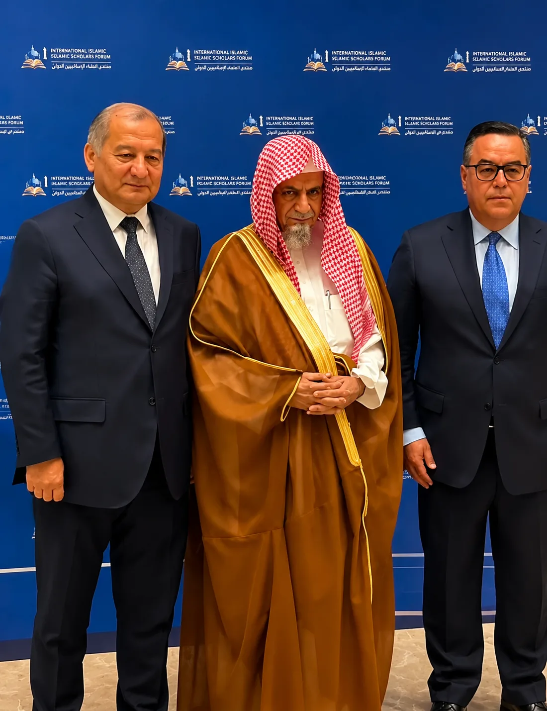 Three men in formal attire pose for a photo at a backdrop with Islamic scholars forum logos; the center man wears a traditional brown thobe and red-checked ghutra with a gold-trimmed bisht.