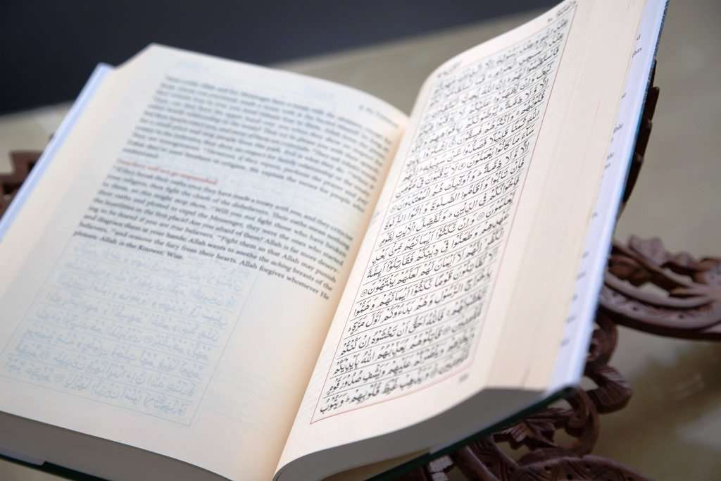 What is the Quran - Nayi Soch News-e-Hub Open book shows dense Arabic script on the right page with notes on the left, resting on a carved stand.