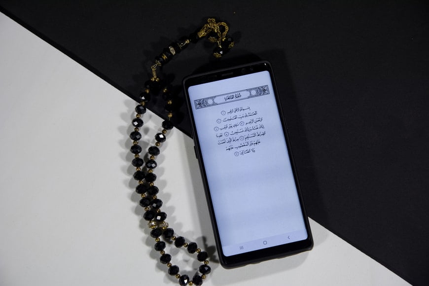 Smartphone displaying Arabic text on screen, placed beside a black and gold bead tasbih on a two-tone white and black background.