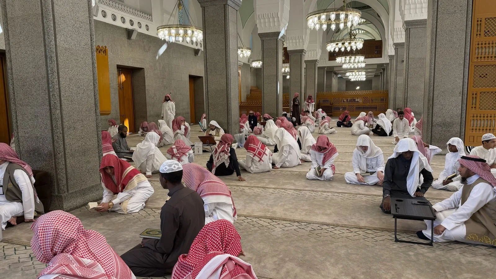 Group of people in white robes and red headscarves sitting on a marble floor in a mosque, reading scriptures among columns and chandeliers.